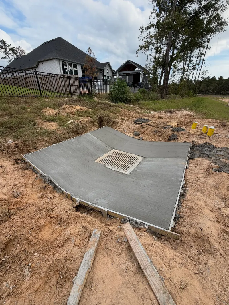 Concrete pad with a drain grate set in the middle, houses behind a fence in the background