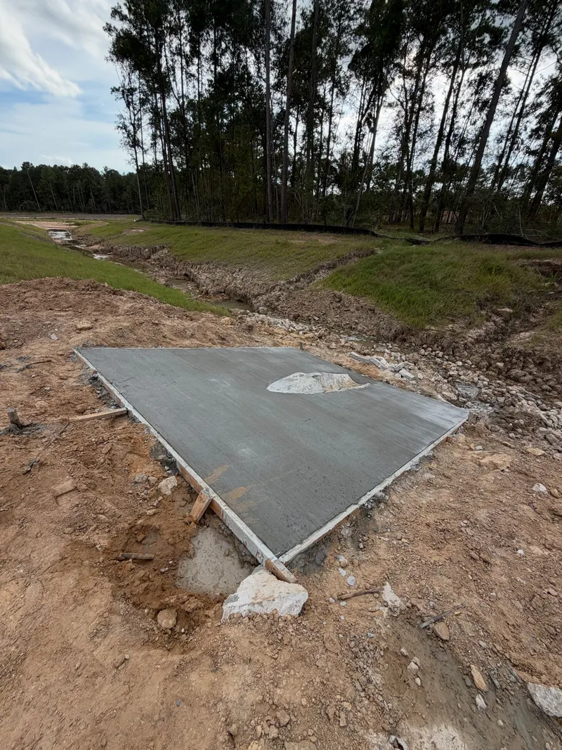 New square concrete pad on cleared ground beside a drainage ditch and a stand of pine trees