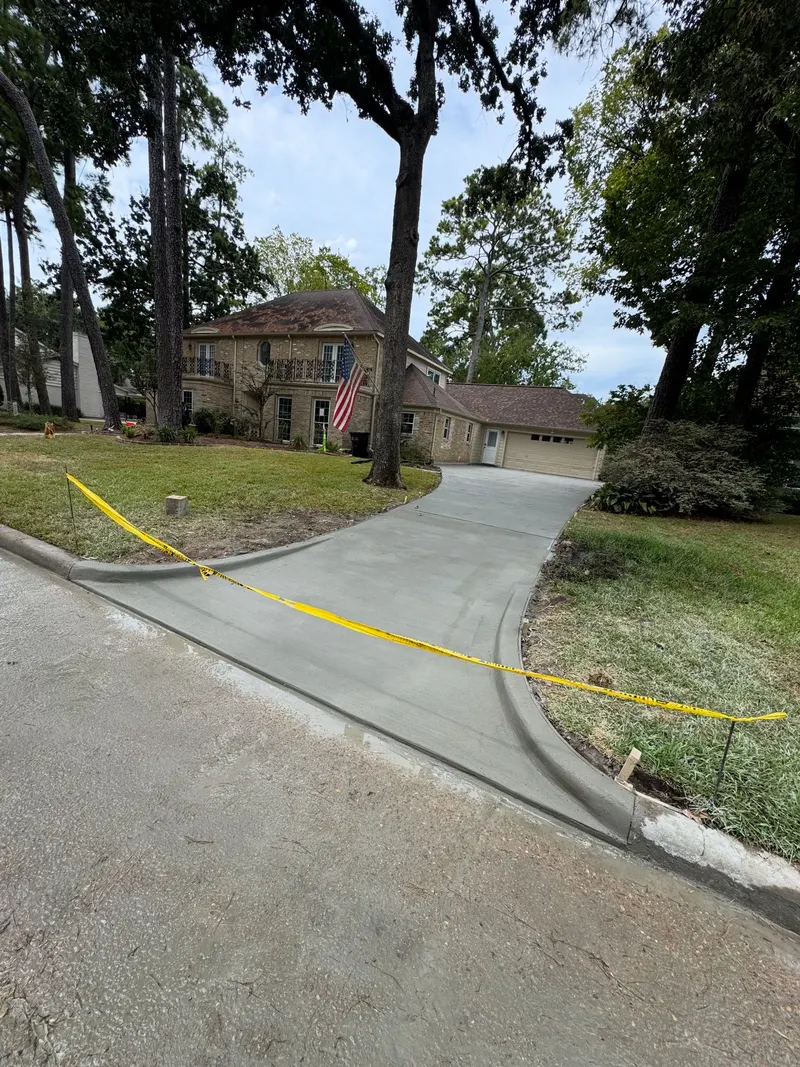 New concrete driveway curving up to a brick house under tall trees, caution tape along the edge