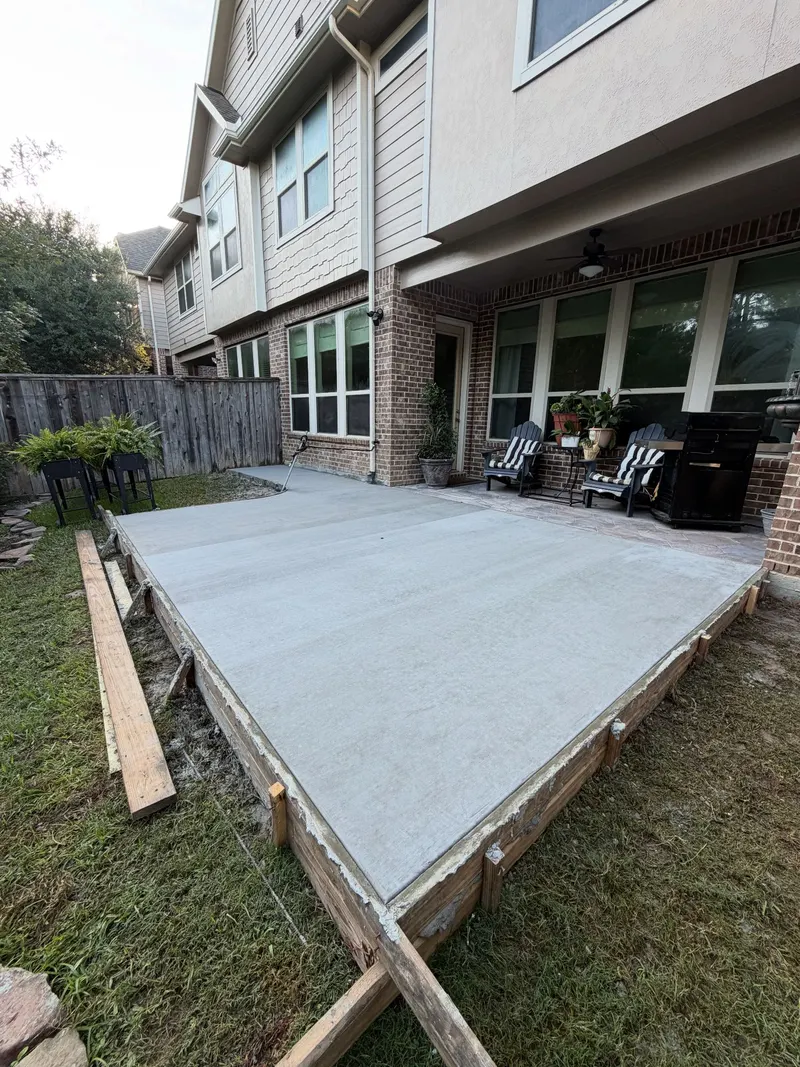 Fresh concrete patio inside wood forms behind a two-story townhome, patio chairs on the porch